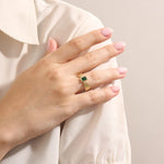 A model wears a vintage-inspired 14k gold statement ring with a bezel-set emerald cut green gemstone. The bold rectangular ring sits comfortably on the finger, catching the light with its glossy finish.