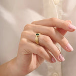 A model wears a vintage-inspired 14k gold statement ring with a bezel-set emerald cut green gemstone. The bold rectangular ring sits comfortably on the finger, catching the light with its glossy finish.