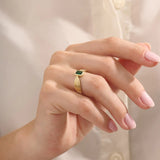 A model wears a vintage-inspired 14k gold statement ring with a bezel-set emerald cut green gemstone. The bold rectangular ring sits comfortably on the finger, catching the light with its glossy finish.