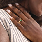 A model wears a vintage-inspired 14k gold statement ring with a bezel-set emerald cut green gemstone. The bold rectangular ring sits comfortably on the finger, catching the light with its glossy finish.