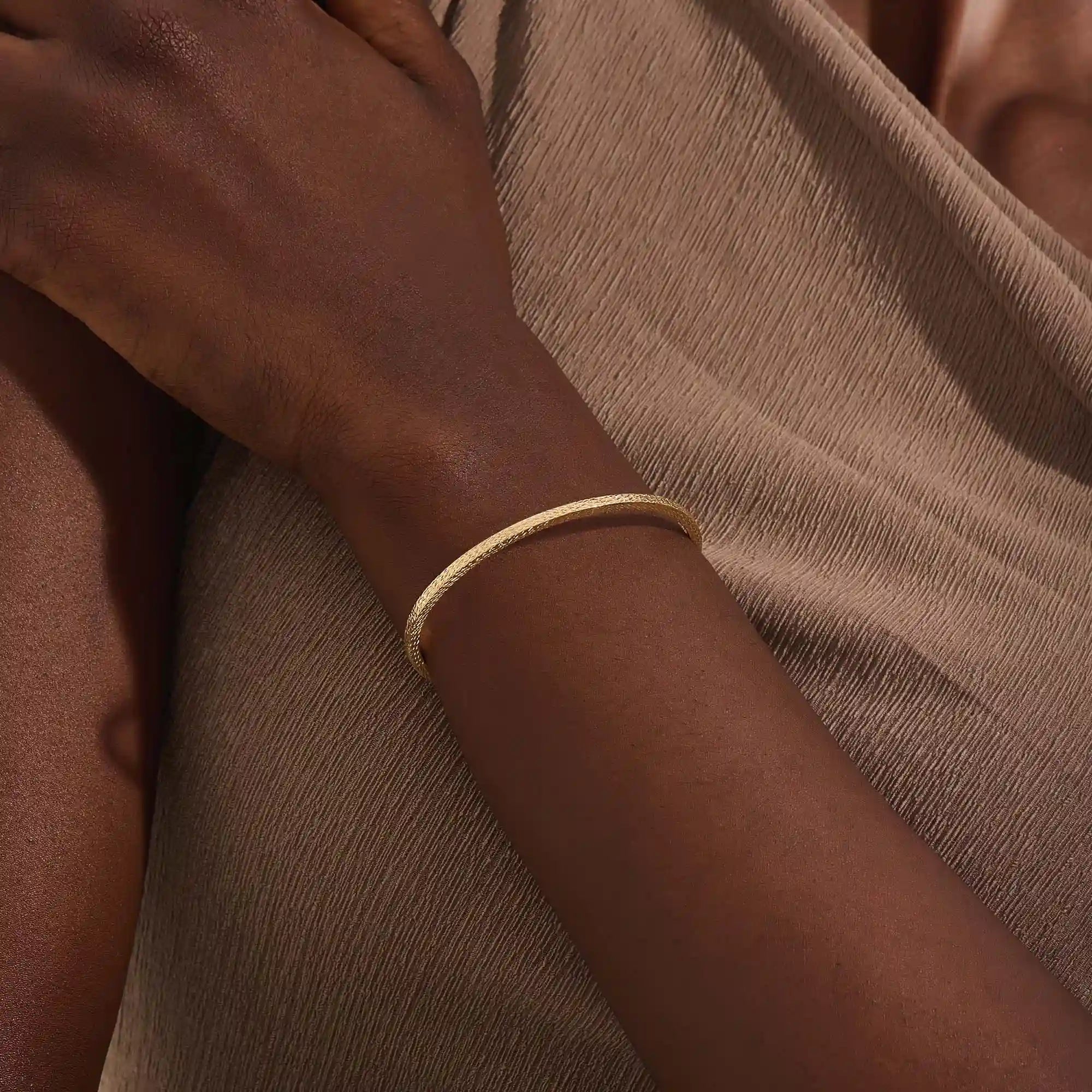 Close-up of a model’s wrist wearing a 14k yellow gold textured square tube bangle with a slim geometric band and polished finish.
