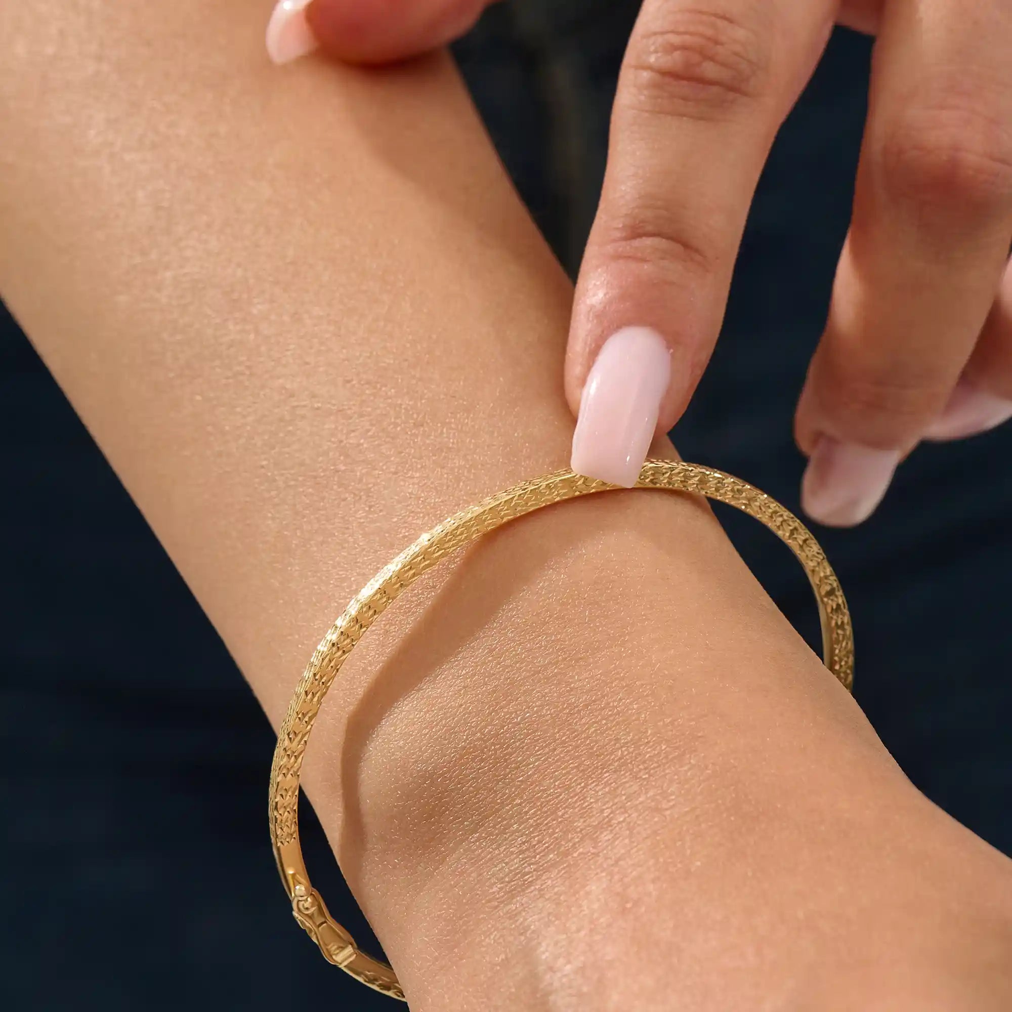 Close-up of a model’s wrist wearing a 14k yellow gold textured square tube bangle with a slim geometric band and polished finish.