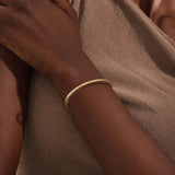 Close-up of a model’s wrist wearing a 14k yellow gold textured square tube bangle with a slim geometric band and polished finish.
