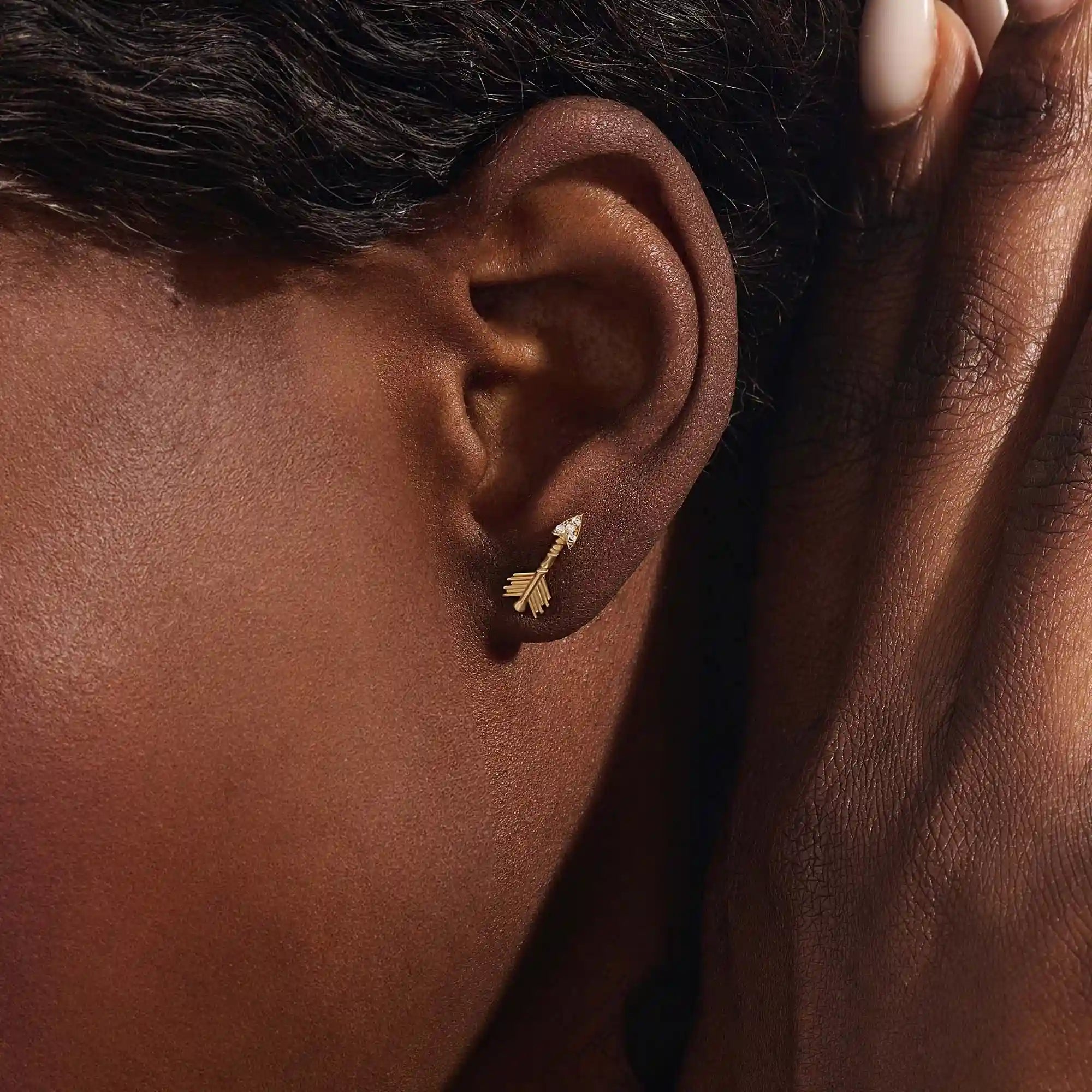 Close-up of a model wearing a 14k solid yellow gold arrow stud earring, showing the sparkle of moissanite stones in the arrowhead.
