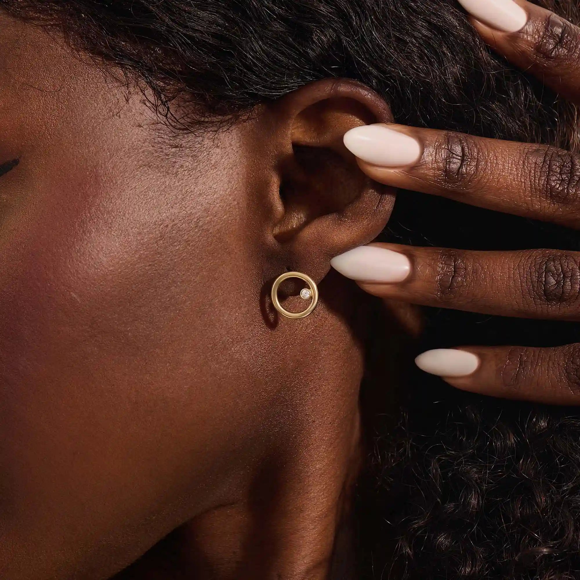 Close-up of a model wearing a 14k yellow gold karma circle stud with a single bezel-set moissanite detail.
