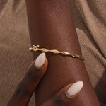 Close-up of a woman wearing a 14k solid yellow gold twist bangle bracelet, showing the bracelet’s sculpted wave design glimmering in natural light.
