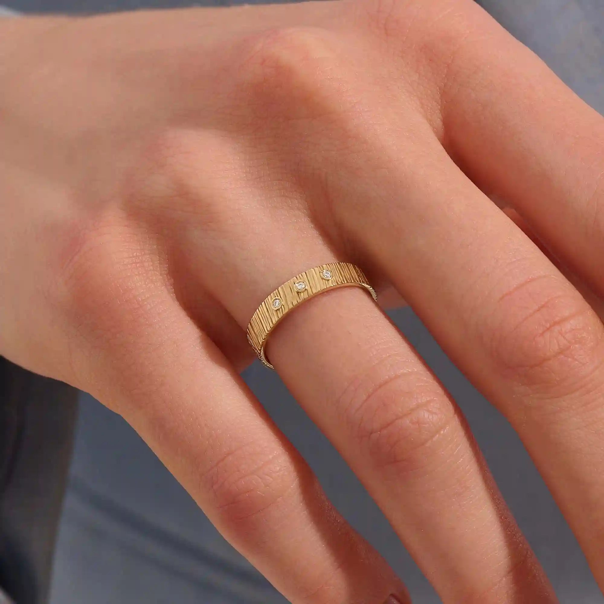 Close-up of a woman wearing a 14k solid yellow gold wide wedding band with wood grain texture and three bezel set round brilliant cut moissanite stones, distinctive and stylish statement jewelry
