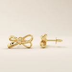 Close-up of simple ribbon stud earrings in polished 14k gold featuring a graceful bow silhouette.
