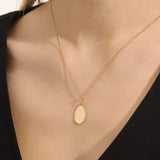 Model wears an oval plain pendant necklace in 14k gold, ideal for personal engravings. A sleek and versatile everyday accessory.