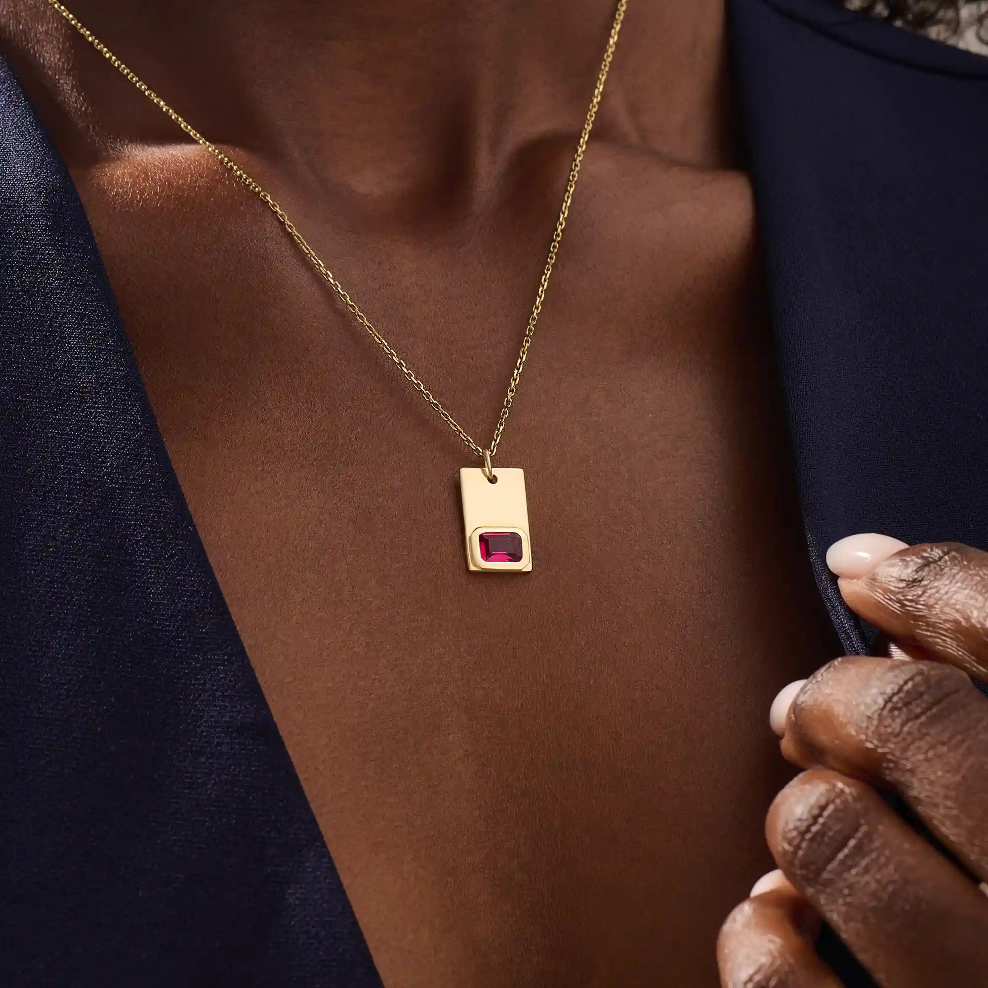 Woman wearing a 14k solid gold rectangle pendant necklace featuring an emerald-cut lab-created ruby in bezel setting. Striking birthstone jewelry with timeless elegance. Ideal for everyday wear or special occasions.